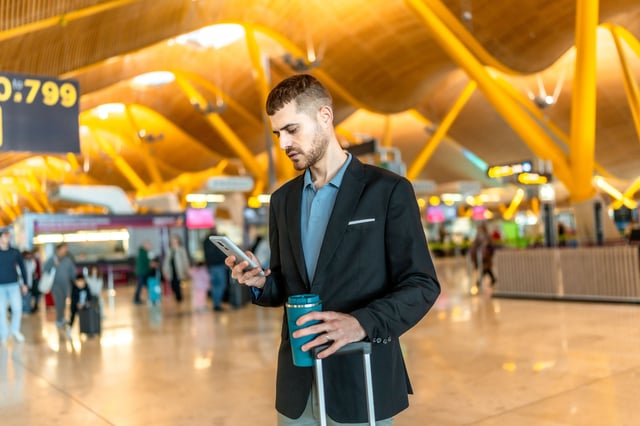 นักธุรกิจรอเที่ยวบินโดยใช้สมาร์ทโฟนและถือแก้วกาแฟแบบใช้ซ้ำได้ที่สนามบิน นักธุรกิจรอเที่ยวบินโดยใช้สมาร์ทโฟนและถือแก้วกาแฟแบบใช้ซ้ำได้ที่สนามบิน