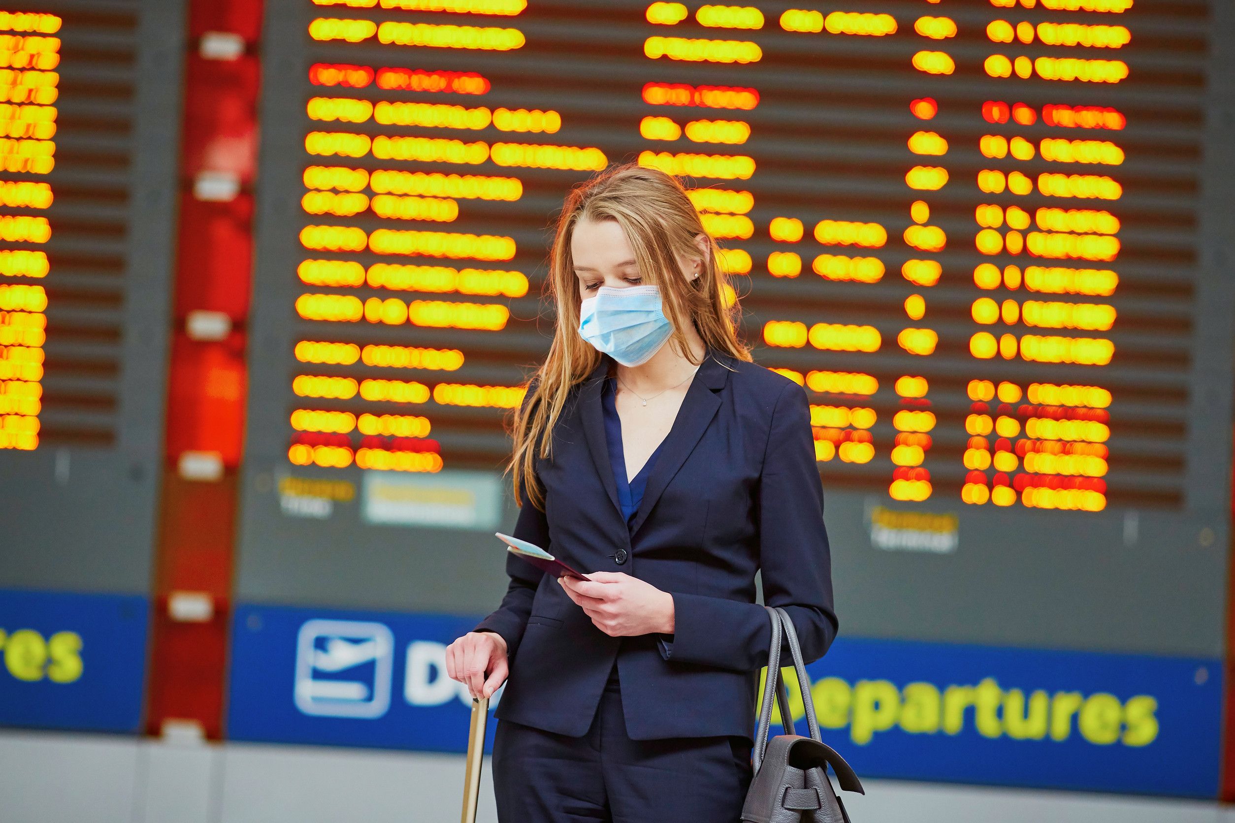  woman wearing protective face mask with hand luggage in international airport terminal looking