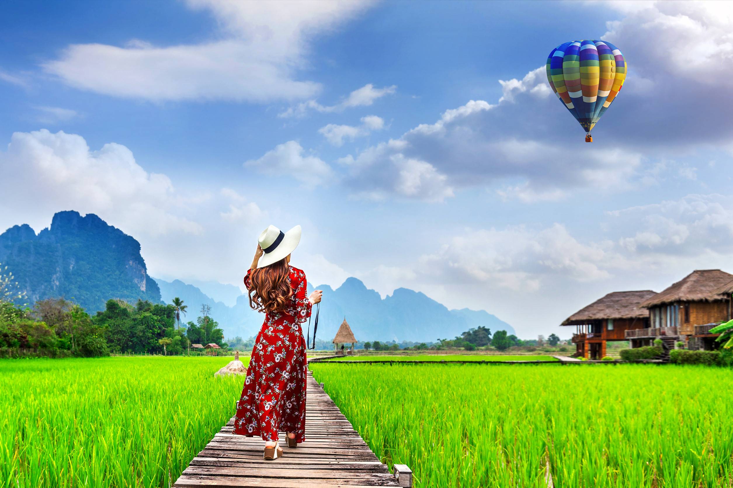 Young woman looking at a balloon and walking on a wooden path with green rice fields in Vang Vieng, Laos